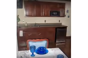 A small kitchen area with wooden cabinets, a countertop, a microwave, a mini refrigerator, and a sink. In the foreground, there is a dining table set with blue glassware and a white tablecloth. A chair with a cushioned backrest is partially visible.