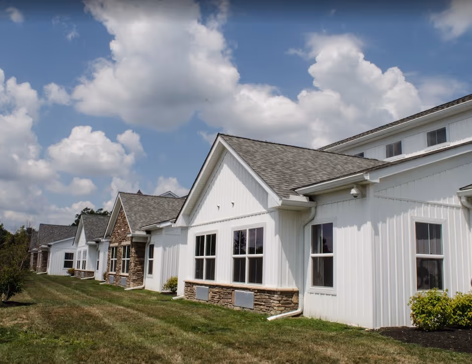 Exterior view of a white building with multiple peaked roofs and large windows, surrounded by green grass and bushes under a partly cloudy blue sky.