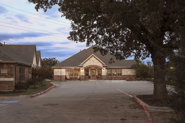 Exterior view of a single-story brick and stone building with a peaked roof, surrounded by trees and a parking lot with marked spaces, under a cloudy sky at dusk.