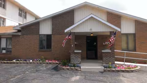 Brick building entrance of Blueberry Hill Assisted Living with flags and flower beds.