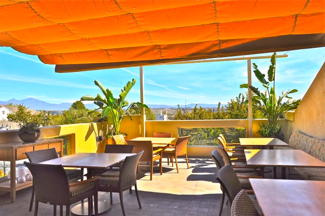Outdoor patio area at The Covington with several tables and wicker chairs arranged for dining or socializing. The space is shaded by an orange awning and decorated with large potted plants. In the background, there is a view of trees and distant mountains under a clear blue sky.