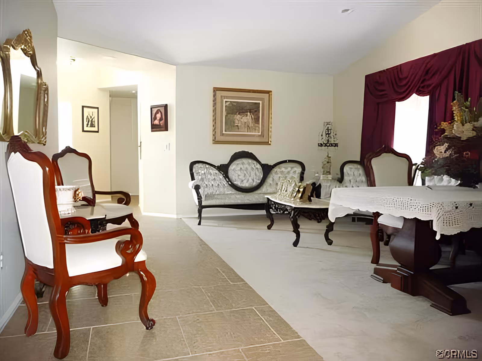 A living room with vintage-style furniture including a white and dark wood armchair, a matching sofa, a coffee table, and a dining table with a white lace tablecloth. The room has light-colored walls, a large window with dark red curtains, framed artwork on the walls, and a decorative mirror.