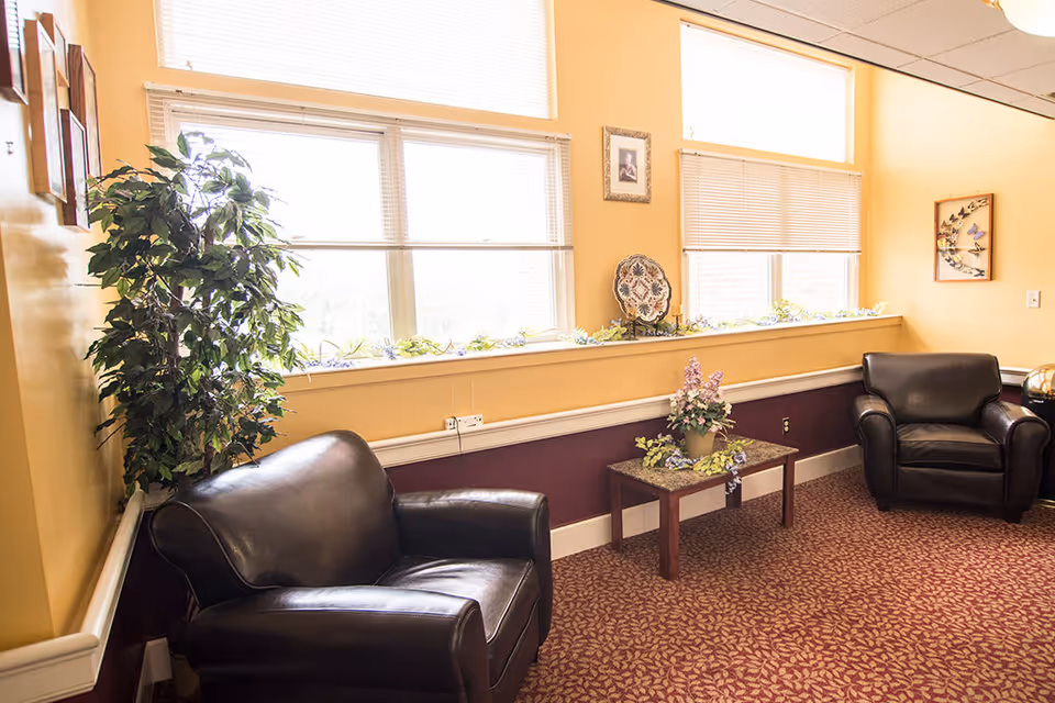A cozy sitting area with two black leather armchairs positioned near large windows with blinds. A small wooden table with a floral arrangement sits between the chairs. The walls are painted yellow and maroon, decorated with framed artwork and a leafy green plant in the corner. The floor is covered with a patterned carpet.