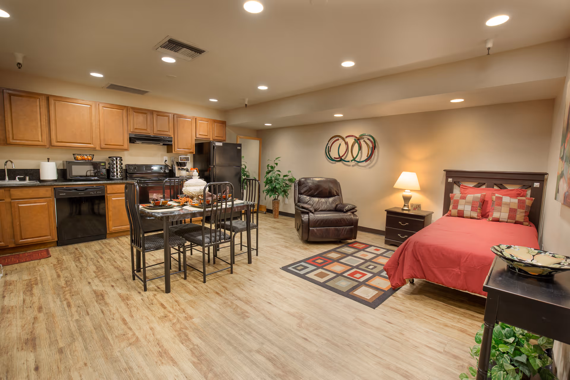 A spacious studio apartment featuring a kitchen area with wooden cabinets, black appliances including a refrigerator, stove, microwave, and dishwasher. In the center, there is a dining table set with four chairs and place settings. To the right, a cozy living area includes a brown leather recliner, a nightstand with a lamp, and a bed with red bedding and decorative pillows. The room has wood flooring, recessed lighting, and decorative wall art.