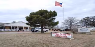 Single-story nursing and rehabilitation facility exterior with American flag, trees, and signage.