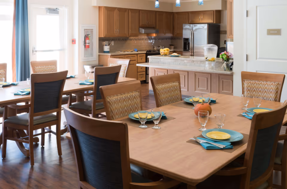 A dining area in a senior living facility with wooden tables and chairs set with yellow plates, blue napkins, and glassware. In the background, there is a kitchen with wooden cabinets, a stainless steel refrigerator, and a countertop with a glass pitcher of water and a bowl of lemons. A door labeled 'Janitor' is visible on the right side.