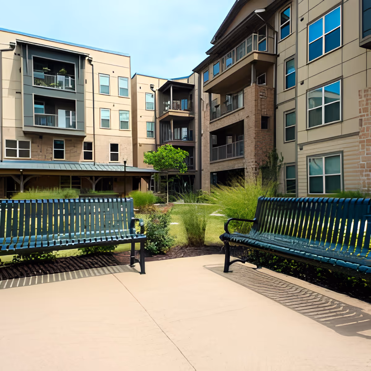 Outdoor courtyard area of a senior living facility with two green metal benches facing each other, surrounded by landscaped greenery and multi-story beige buildings with balconies and windows under a clear blue sky.