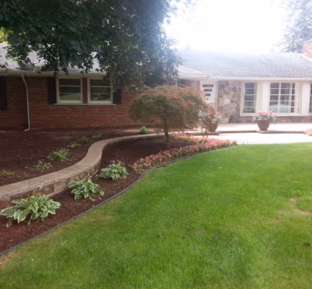 Front view of a single-story brick house with a curved landscaped flower bed, potted plants, and a green lawn.