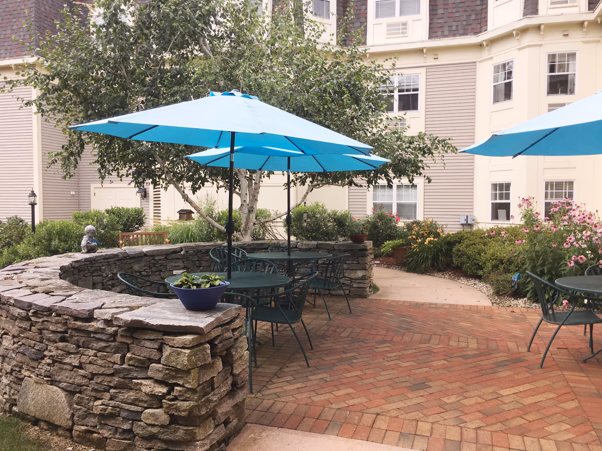 Outdoor patio area at Middlesex Health at One MacDonough Place featuring round metal tables with blue umbrellas, surrounded by metal chairs. The patio is paved with red bricks in a herringbone pattern and bordered by a low stone wall. There are green bushes, flowering plants, and a tree providing additional shade. The background shows the exterior of a beige and tan building with multiple windows.