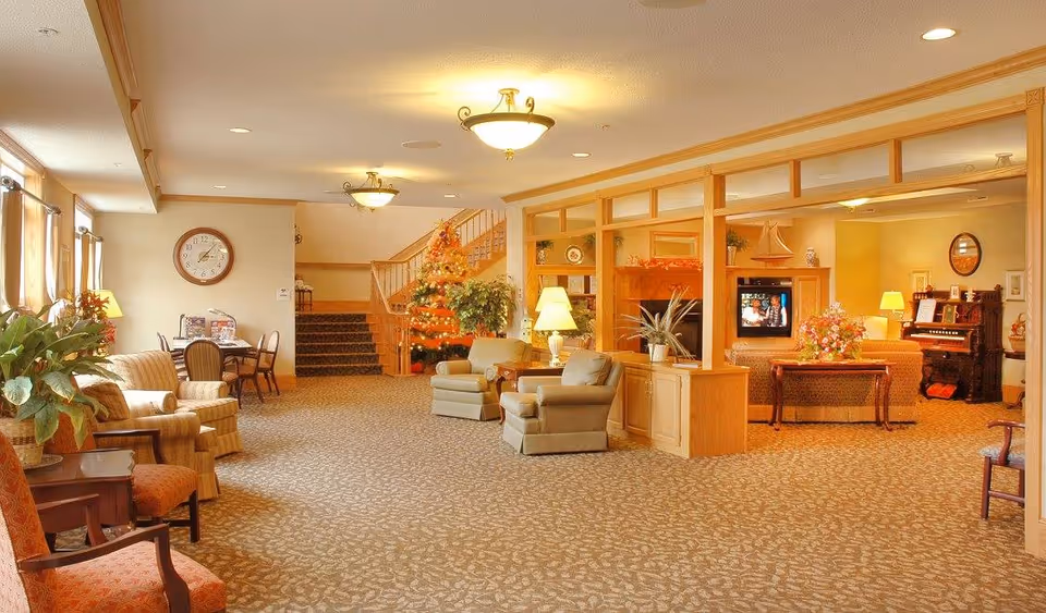 A spacious and warmly lit senior living facility common area with comfortable armchairs, a Christmas tree decorated with lights, a clock on the wall, a television, and a piano. The room features beige walls, patterned carpet, wooden trim, and large windows with curtains letting in natural light.