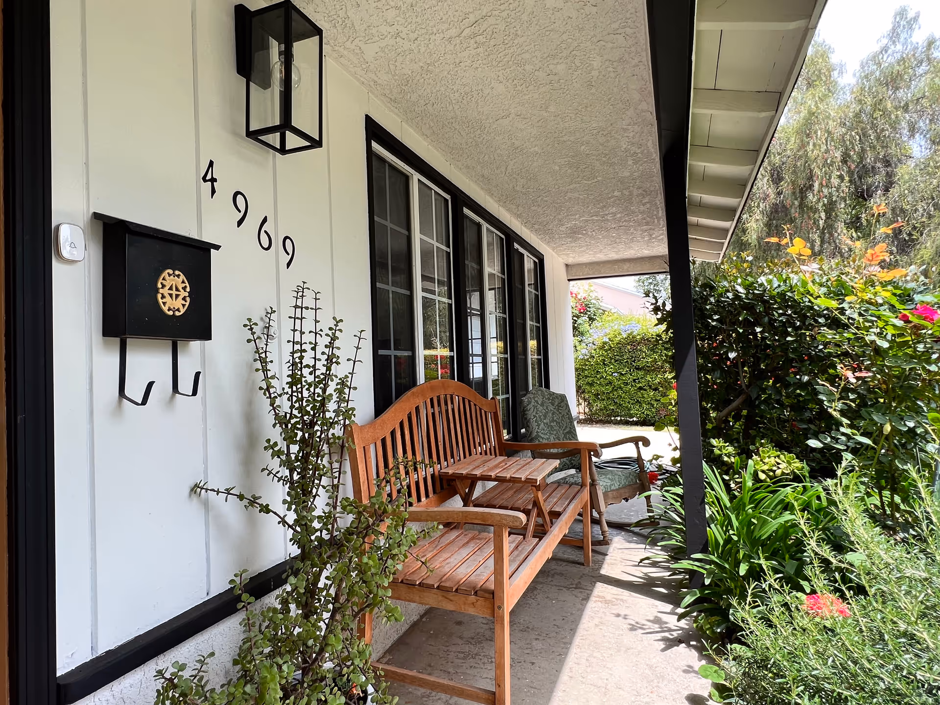 Front porch area of a senior living facility with a wooden bench, a cushioned armchair, a small wooden table, a black mailbox with a decorative emblem, house number 4969 on the white wall, a black-framed window, a black outdoor light fixture, and lush green plants and bushes along the walkway.