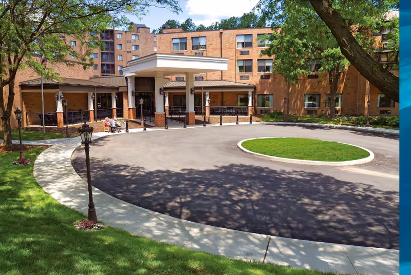 Front exterior view of a multi-story brick building with a covered entrance and circular driveway surrounded by green grass and trees. Two people are sitting on a bench near the entrance.