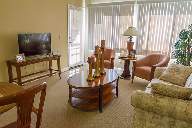 A cozy living room with a floral patterned sofa, two pink armchairs, a wooden coffee table with decorative candle holders, a side table with a lamp and framed photo, a flat-screen TV on a wooden stand, and large windows with vertical blinds letting in natural light.