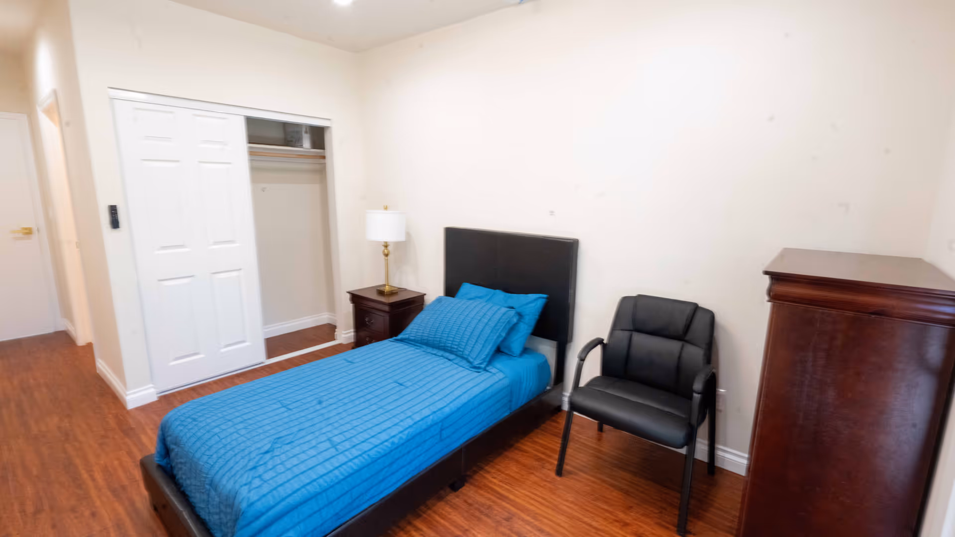 A simple bedroom with a single bed covered in a blue bedspread and matching pillows. Next to the bed is a dark wooden nightstand with a lamp on it. There is a black padded chair beside the bed and a tall dark wooden dresser against the wall. The room has wooden flooring, white walls, and a closet with sliding white doors.