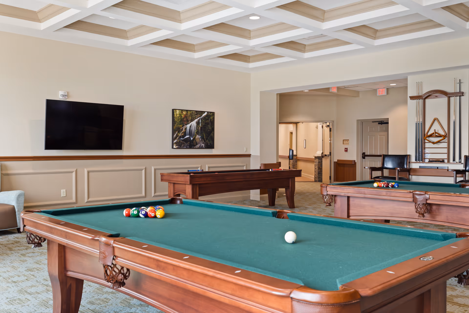 A recreational room with two pool tables, one in the foreground with billiard balls arranged and a cue ball, and another in the background. The room has a coffered ceiling, a wall-mounted flat screen TV, a framed picture of a waterfall, and a rack with pool cues and triangle racks. There is a chair near the pool cue rack and an open doorway leading to a hallway.