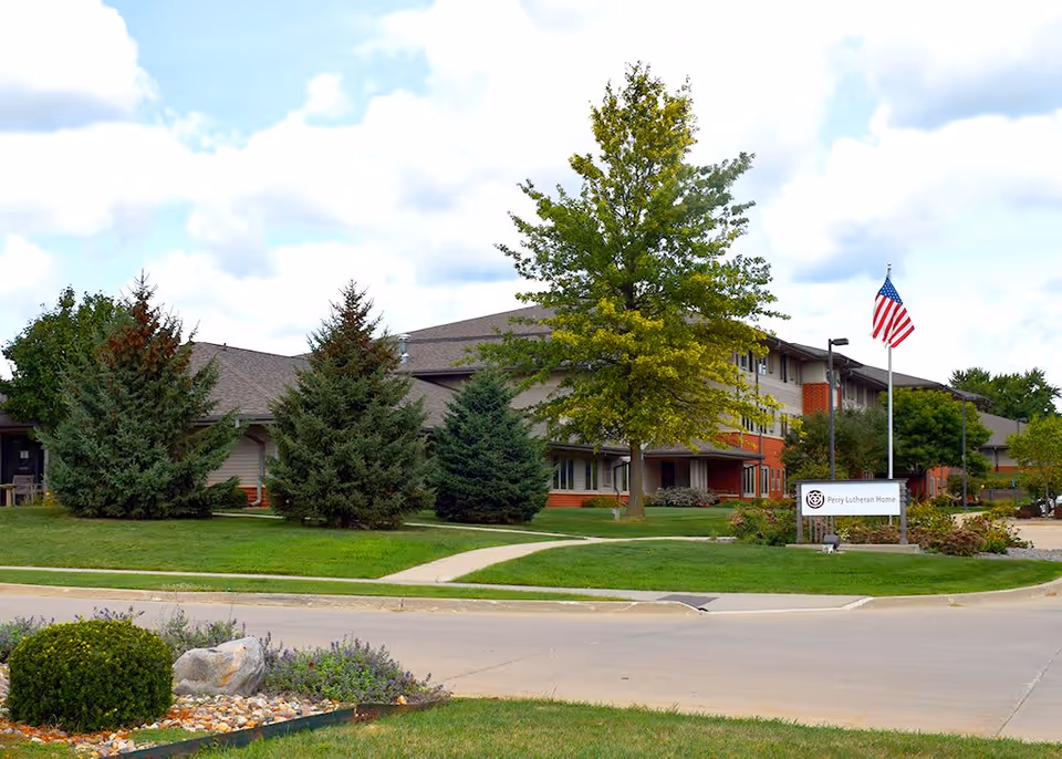 Exterior view of the Spring Valley Campus of Perry Lutheran Homes, showing a multi-story building surrounded by green trees and well-maintained lawns. An American flag is flying on a flagpole near a sign that reads 'Perry Lutheran Home'. The sky is partly cloudy.
