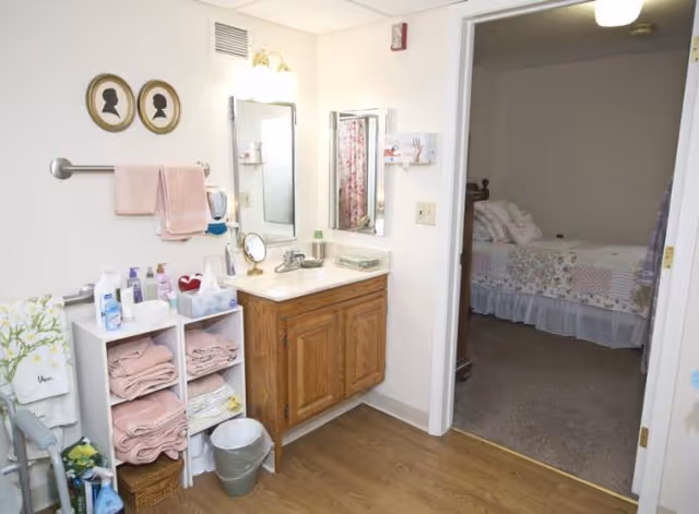 A small bathroom with a wooden vanity and sink, shelving holding folded towels and toiletries, and an open door showing a bed in the adjacent bedroom.