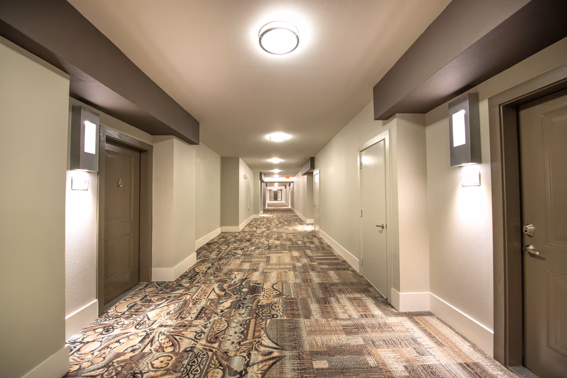 Long indoor hallway in a senior living facility with patterned carpet, beige walls, multiple closed doors on both sides, and ceiling lights evenly spaced along the corridor.