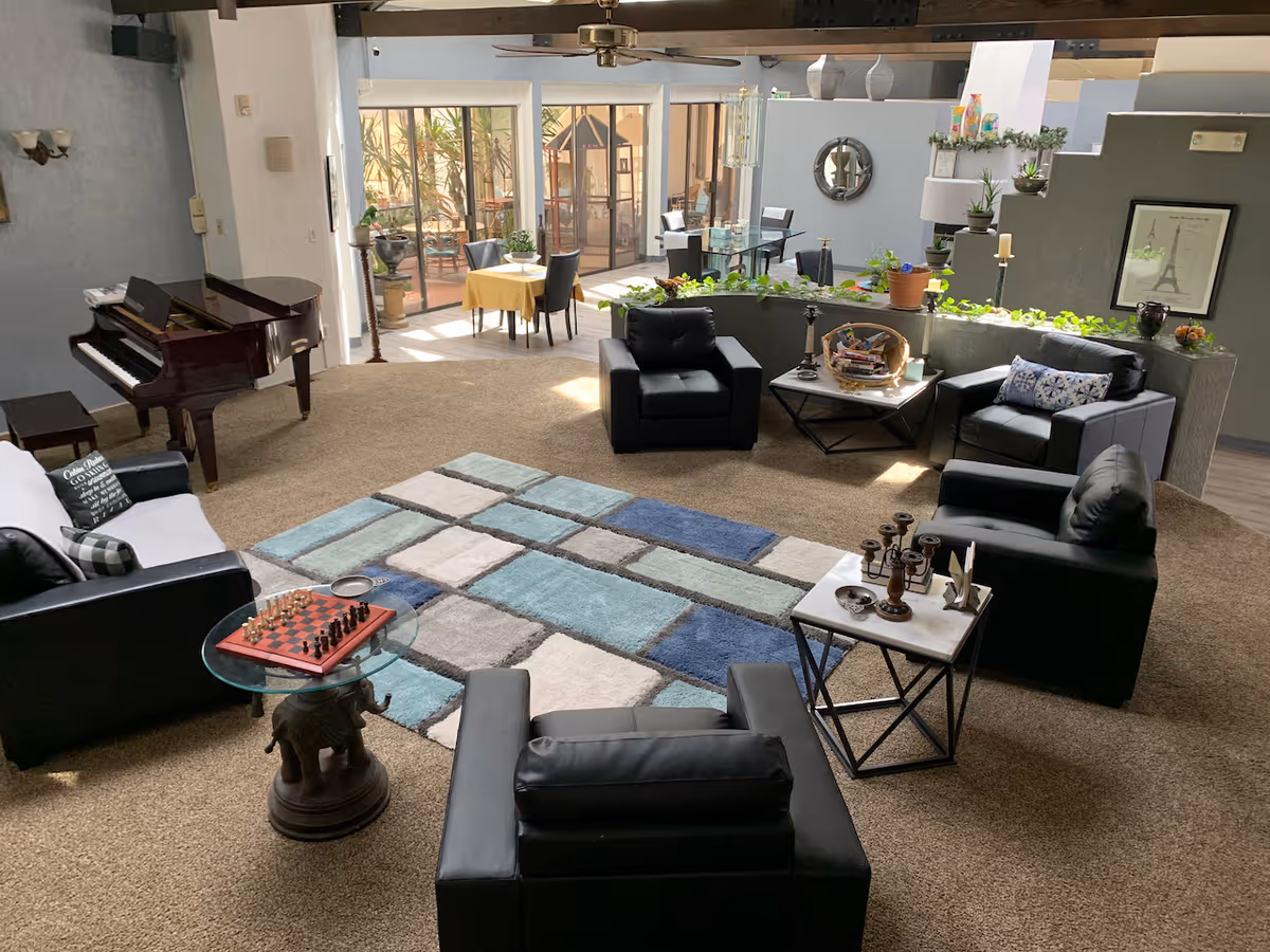 A spacious living room area with a beige carpet and a multicolored geometric rug in the center. The room features four black leather armchairs arranged around a small coffee table with books and decorative items. There is a black and white sofa with pillows, a glass side table with a chessboard, and a grand piano against the wall. In the background, there are dining tables with chairs near large windows that let in natural light, and various plants and decorative items are placed around the room.