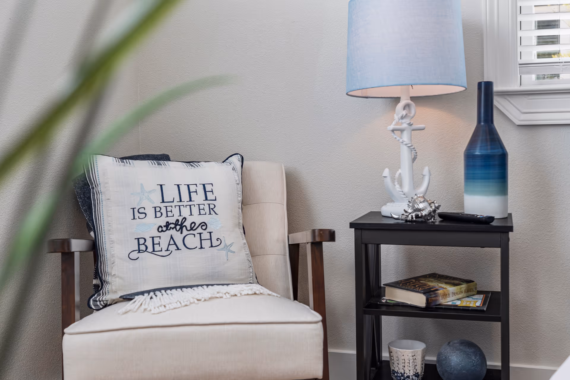A cozy corner with a beige armchair featuring a decorative pillow that reads 'Life is better at the beach.' Next to the chair is a black side table holding a white anchor-themed lamp with a blue lampshade, a blue and white gradient vase, a remote control, a seashell decoration, and some books. A window with white blinds is partially visible in the background.