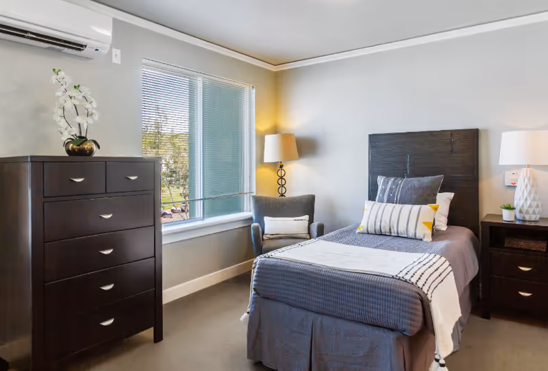 A neatly arranged bedroom with a single bed covered in gray bedding and decorative pillows. To the right of the bed is a dark wooden nightstand with a white lamp and a small plant. To the left of the bed is a gray armchair with a cushion and a tall floor lamp behind it. A large window with blinds lets in natural light. On the left side of the room is a tall dark wooden dresser with a white orchid plant on top. The walls are painted light gray and the room has a calm, cozy atmosphere.