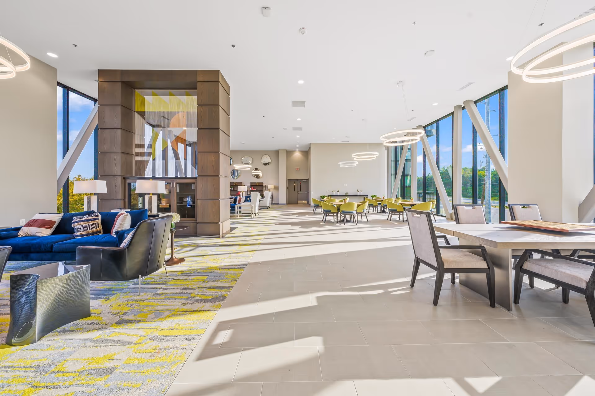 Bright and spacious common area with large floor-to-ceiling windows allowing natural light to fill the room. The space features modern furniture including blue sofas, black armchairs, green chairs around tables, and a large decorative fireplace in the center. Circular pendant lights hang from the ceiling, and the floor is a combination of patterned carpet and light-colored tiles.