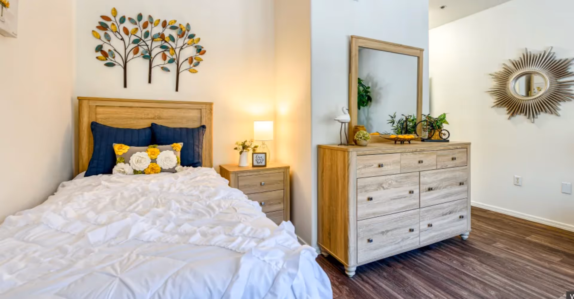 Bright bedroom with a single bed dressed in white, decorative pillows and wall art, a bedside table with a lamp, and a wooden dresser with mirror.