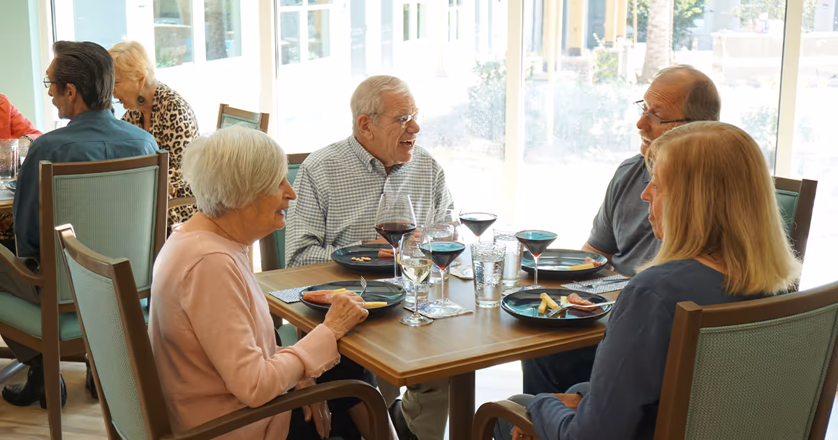 Four elderly people sitting around a dining table in a bright room with large windows, enjoying a meal with plates of food and glasses of wine and water, engaging in conversation.