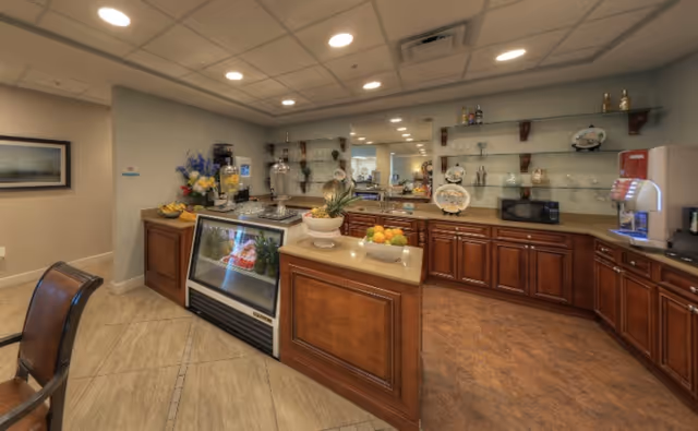 Interior view of a kitchen area with wooden cabinets, a countertop with fruit bowls, a glass-front refrigerator, a microwave, and various kitchen appliances. The room has a tiled floor, recessed ceiling lights, and shelves with decorative plates and glassware.