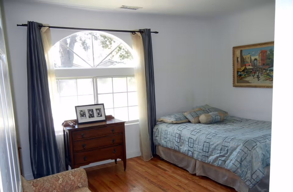 A bedroom with a single bed covered in a blue patterned bedspread and several pillows. There is a wooden chest of drawers under a large window with an arched top, flanked by blue and beige curtains. A framed picture is placed on the chest of drawers. A framed painting hangs on the wall above the bed, and a patterned armchair is partially visible in the foreground. The floor is wooden.
