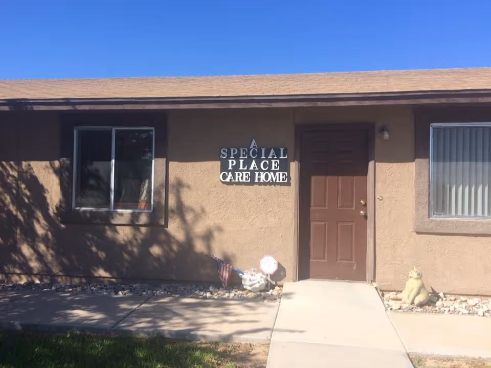 Exterior view of a single-story building with a brown door and two windows. A sign next to the door reads 'A Special Place Care Home'. There is a small American flag and a decorative cat statue near the entrance. The sky is clear and blue.