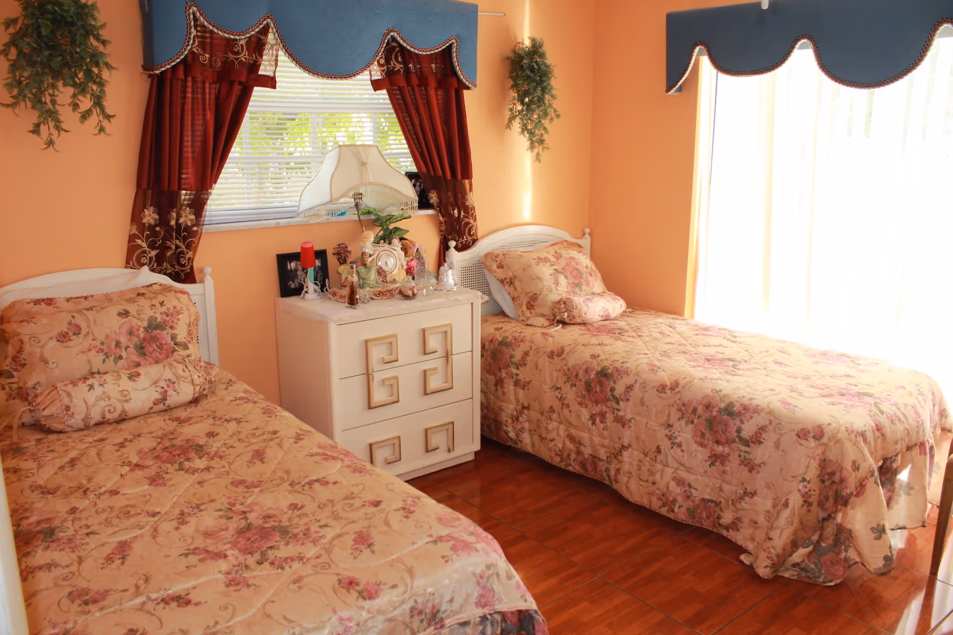 A cozy bedroom with two single beds covered in floral bedspreads. Between the beds is a white nightstand with decorative items including a lamp, framed photos, and small figurines. The walls are painted peach, and the windows have red and blue valances with curtains. There are hanging plants on the walls and wooden flooring.