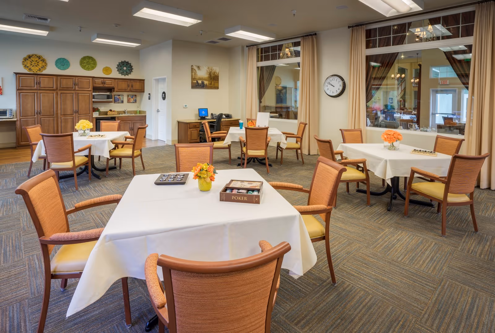 A bright and spacious common area with several tables covered in white tablecloths, each surrounded by wooden chairs with orange cushions. The tables have small flower arrangements and board games on them. In the background, there is a kitchenette area with wooden cabinets and a microwave, as well as a desk with a computer. Large windows with beige curtains separate this room from another dining area visible through the glass. A clock is mounted on the wall.