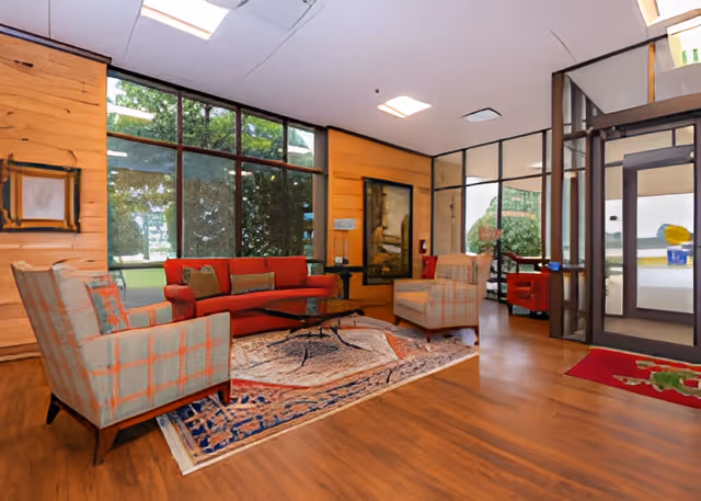 A cozy senior living facility lounge area with a red sofa, two patterned armchairs, a wooden coffee table, and a decorative rug on a wooden floor. Large windows provide a view of greenery outside, and there is a glass door entrance to the right. The walls are wood-paneled with framed artwork.