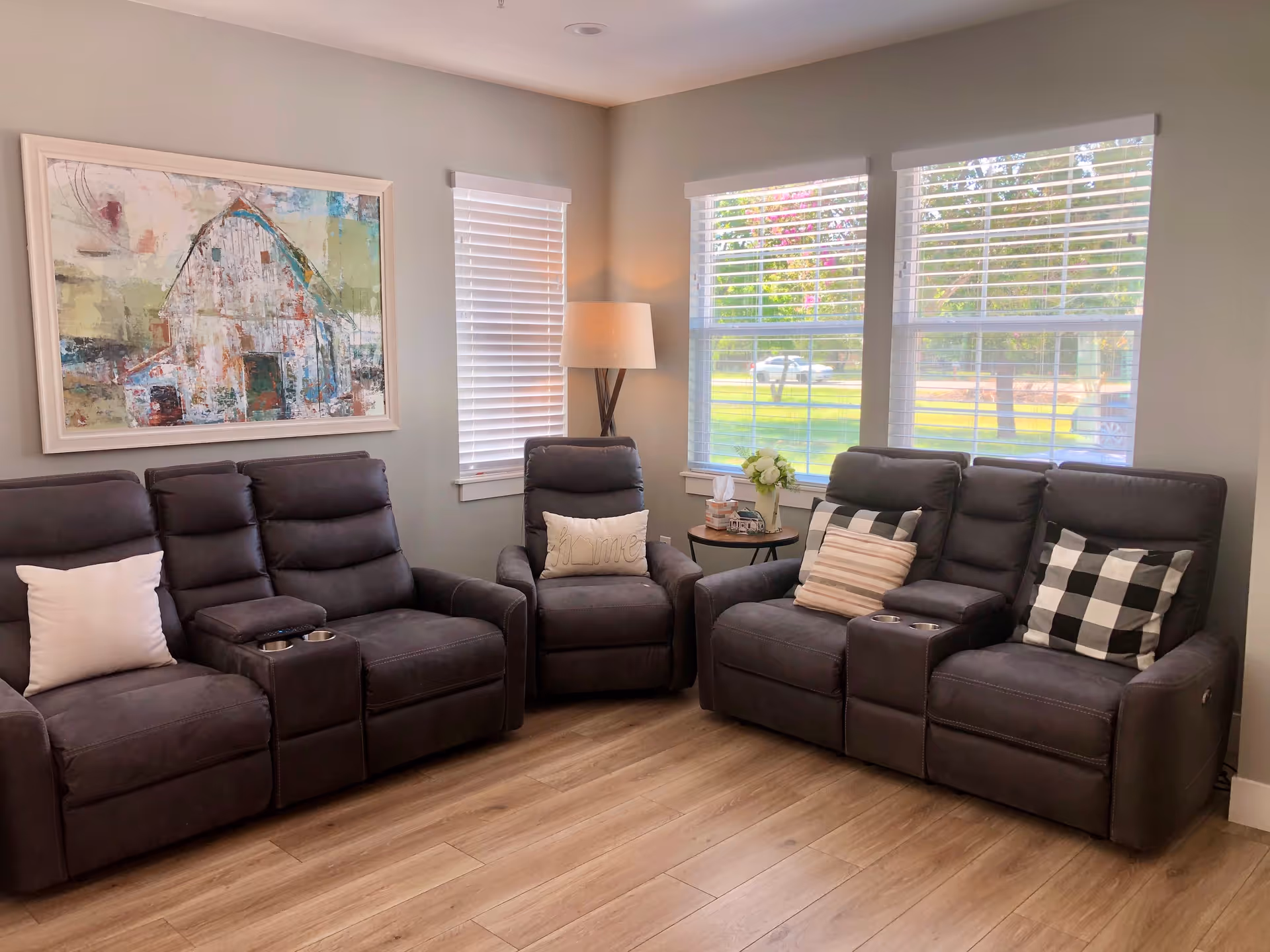 A cozy living room with two dark gray reclining sofas and a matching armchair, each adorned with decorative pillows. The room features light wood flooring, a floor lamp in the corner, and three windows with white blinds letting in natural light. A large framed painting of a barn hangs on the wall above one sofa.