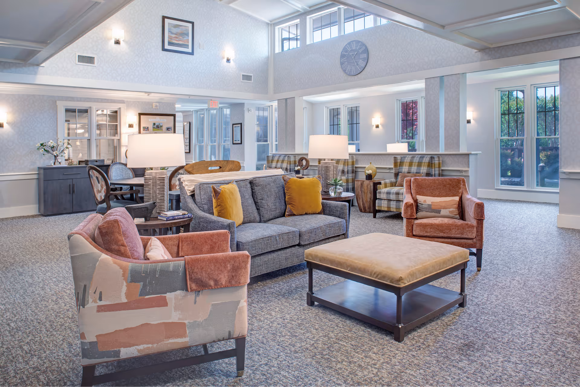 A bright and spacious living room area in a senior living facility with a gray sofa adorned with yellow pillows, two patterned armchairs, two pinkish armchairs, a large cushioned ottoman, and several table lamps. The room features large windows allowing natural light, a wall clock, framed artwork, and a carpeted floor.
