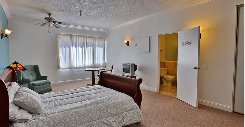 A bedroom in Villa Redondo featuring a large bed with patterned bedding, a green armchair, a small round table with a chair near a window with white curtains, a ceiling fan, and an open door leading to a bathroom with a visible toilet.