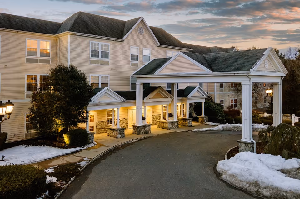 Front entrance of a multi-story beige senior living building with a covered porte-cochere, lit windows, driveway, and patches of snow.