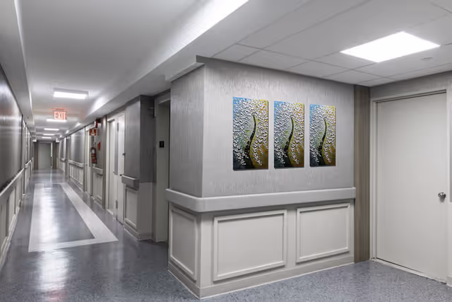 A clean, well-lit hallway in an assisted living facility with light gray walls, a patterned floor, and three abstract art pieces hanging on the wall. There are multiple doors along the hallway and an exit sign at the far end.