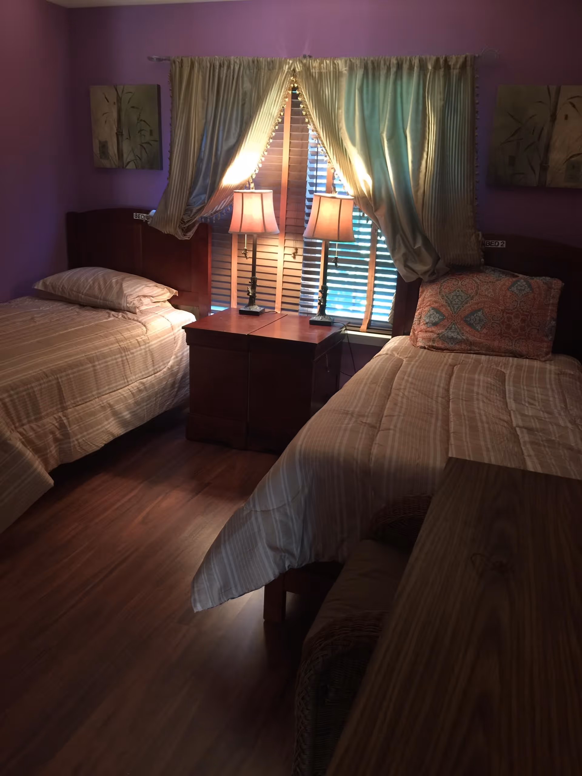 A bedroom with two single beds separated by a wooden nightstand with two lamps. The beds have striped bedding and one pillow has a decorative cover. The room has purple walls, wooden flooring, and a window with cream-colored curtains partially drawn.