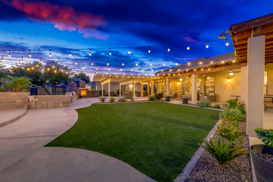 Outdoor patio area at dusk with string lights hanging overhead, a green lawn in the center, seating areas under covered patios, and a fireplace surrounded by stone walls. The sky is vibrant with blue and pink clouds.