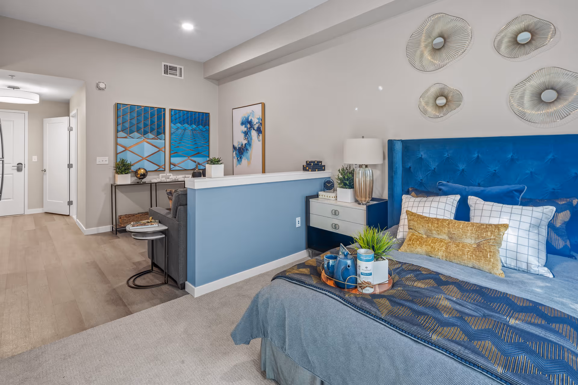 A modern bedroom with a blue upholstered headboard, decorated with multiple pillows in blue, white, and gold. A bedside table with a lamp, books, and a plant is next to the bed. The room features decorative wall art above the bed and a small sitting area with a gray couch and a console table with plants and artwork. The flooring transitions from carpet in the bedroom area to wood in the hallway leading to a white door.