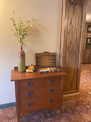 A wooden chest of drawers with three drawers, placed against a wall next to a wooden column. On top of the chest is a tall vase with green and yellow flowers, a small arrangement of decorative gourds, and an open backgammon board with pieces set up. The floor is carpeted with a patterned design, and a wooden cabinet is visible in the background.