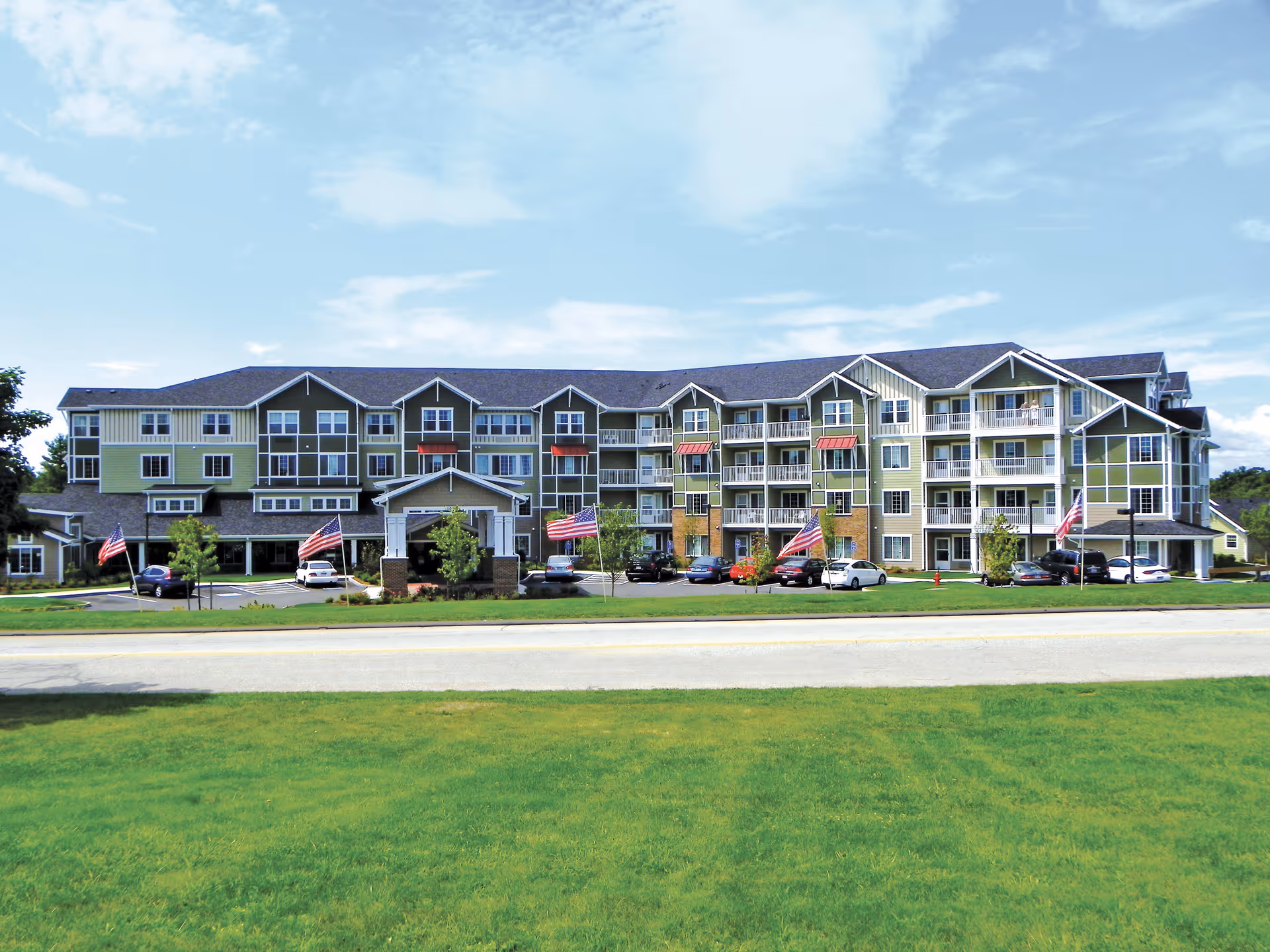 Front exterior of a multi-story senior living building with balconies, a covered entrance, American flags, and a grassy foreground.