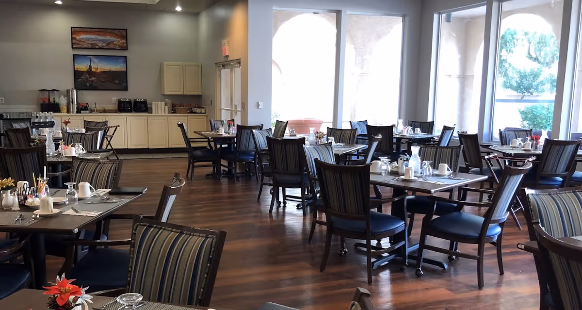 Spacious senior living dining room with multiple set tables and chairs and large windows along one wall.