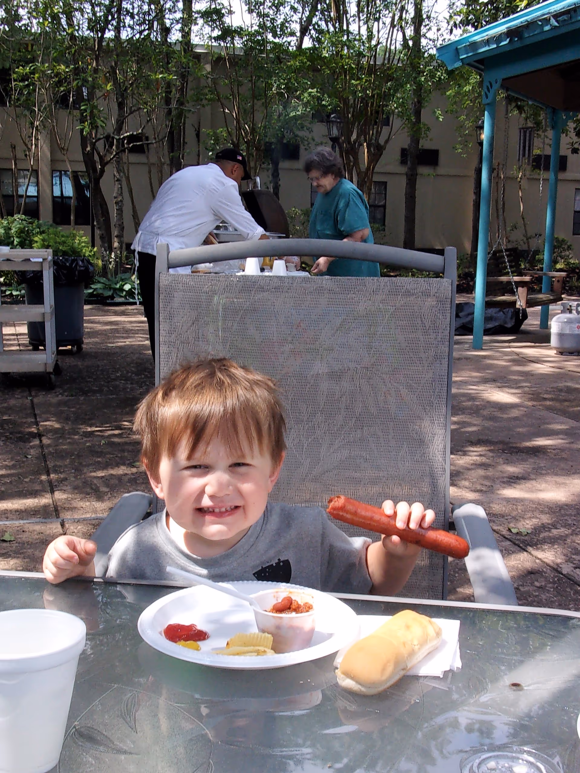 A young boy sitting at an outdoor table holding a hot dog, with a plate of food including chips, ketchup, and a bread roll in front of him. In the background, a man in a white shirt and a woman in a green shirt are standing near a grill in a shaded garden area with trees and a building visible.