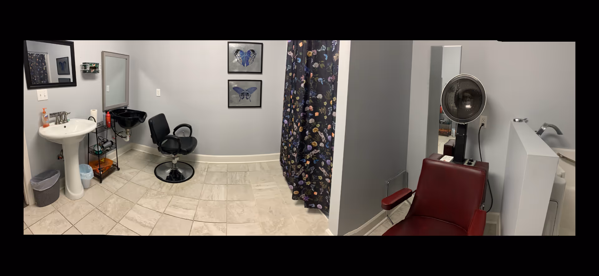 Interior view of a small salon or grooming area in an assisted living facility. The room features a white pedestal sink with a mirror above it on the left side, a black salon chair in front of a black wash basin, and two framed butterfly pictures on the wall. On the right side, there is a maroon salon chair with a vintage hair dryer hood attached. The floor is tiled, and the walls are painted light gray. A floral curtain partially covers a section of the room.