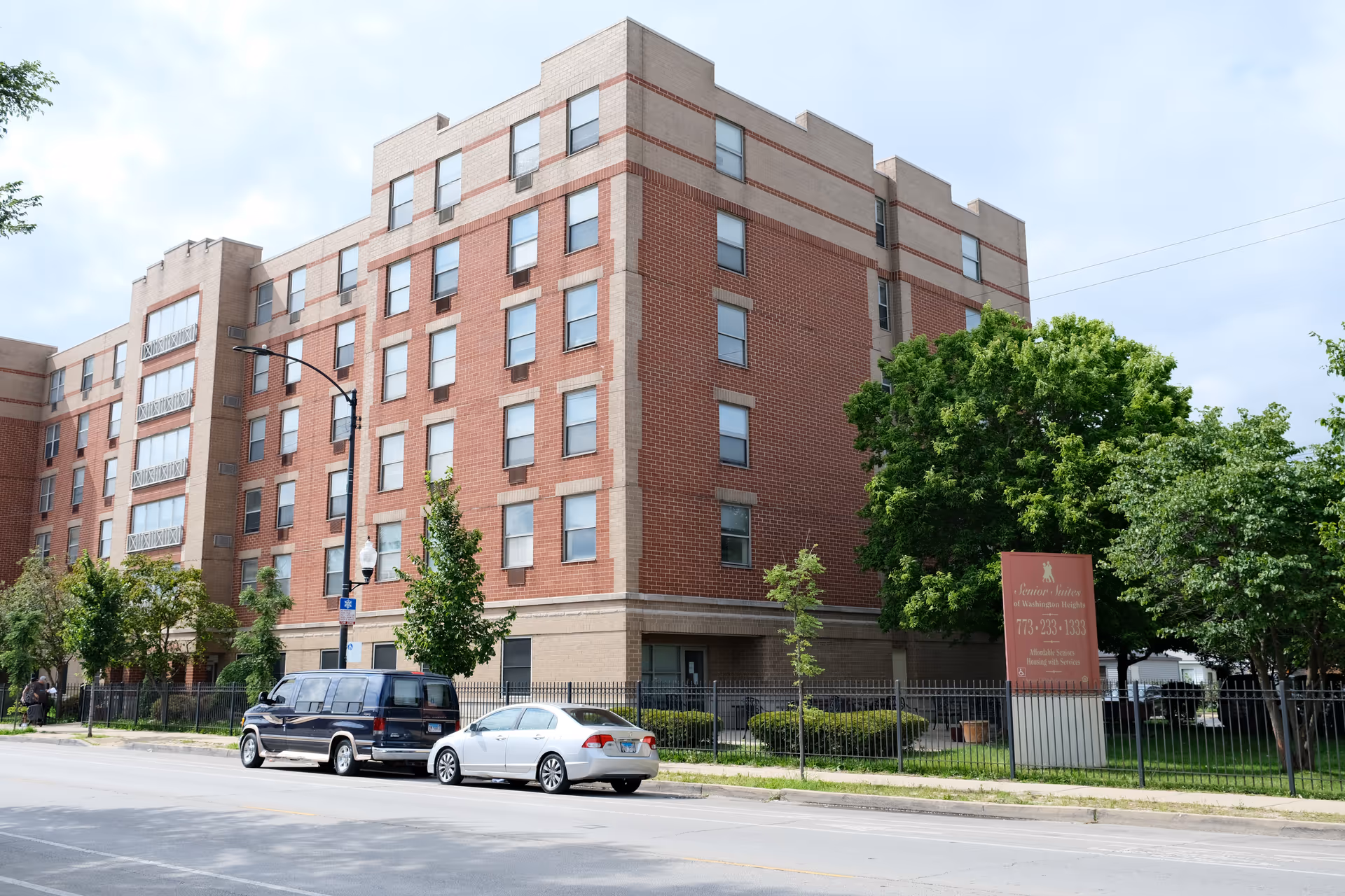 Exterior view of a multi-story brick senior living facility named Senior Suites of Washington Heights with several windows, a fenced yard with trees, and two parked vehicles along the street in front.