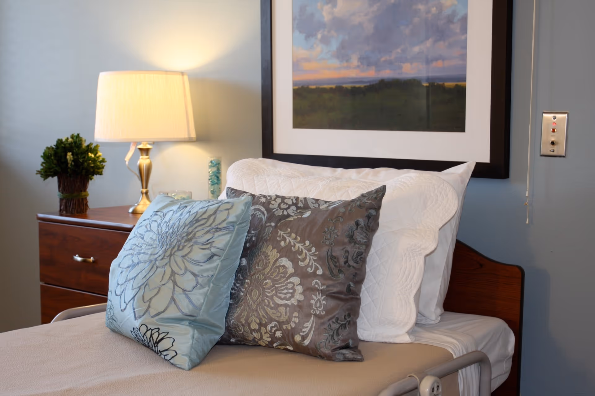 A neatly made bed with white pillows and two decorative cushions, one light blue with a floral pattern and the other gray with a floral design. Next to the bed is a wooden nightstand with a brass lamp, a small green plant, and a glass candle holder. Above the bed hangs a framed landscape painting with a cloudy sky and greenery.