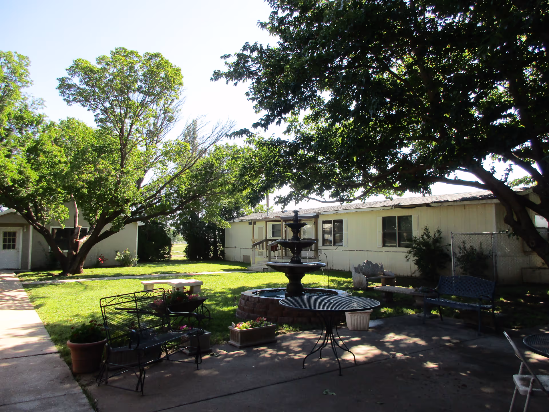 Outdoor courtyard area at Palo Duro Nursing Home featuring a central multi-tiered water fountain surrounded by metal benches and tables. The area is shaded by large trees with green grass and a paved walkway leading to a building in the background.